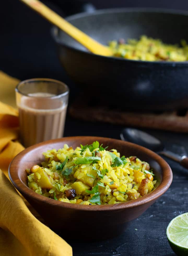 Poha served in a wooden bowl and served with tea. Yellow napkin as well as half a lemon is in the background as well