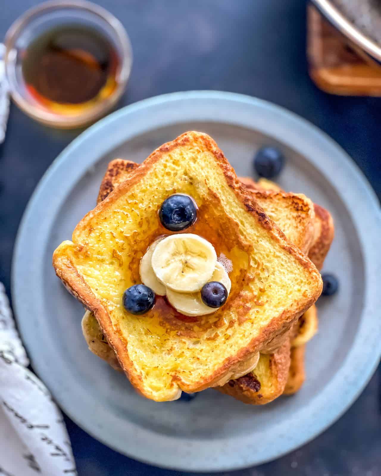 A blue plate of french toast shot from the top down with slices of banana and blueberries on top and a small bowl of syrup to the top left of the plate.