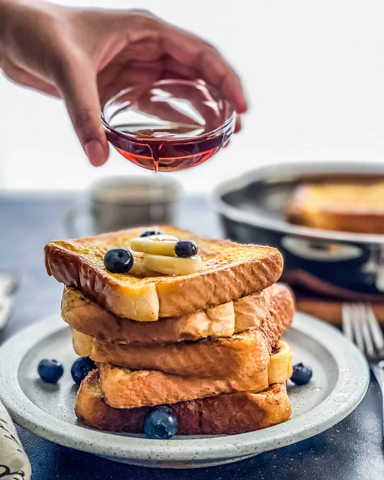 A fresh stack of french toast on a blue plate with a hand pouring syrup on top of the stack.