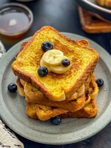 A blue plate with a stack of french toast with blueberries and banana slices on top and a small bowl of syrup in the background.