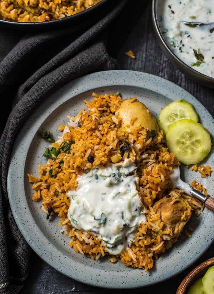 An overshot of biryani served with cucumber raita and a few slices of cucumber on a gray plate