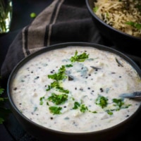 Cucumber raita served in a black bowl accompanied by jeera rice in a black bowl along with a black napkin on the side