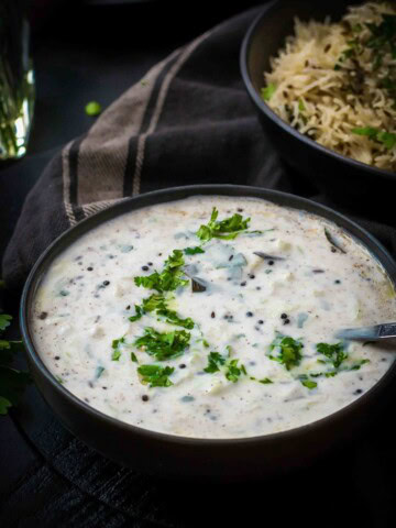 Cucumber raita served in a black bowl accompanied by jeera rice in a black bowl along with a black napkin on the side