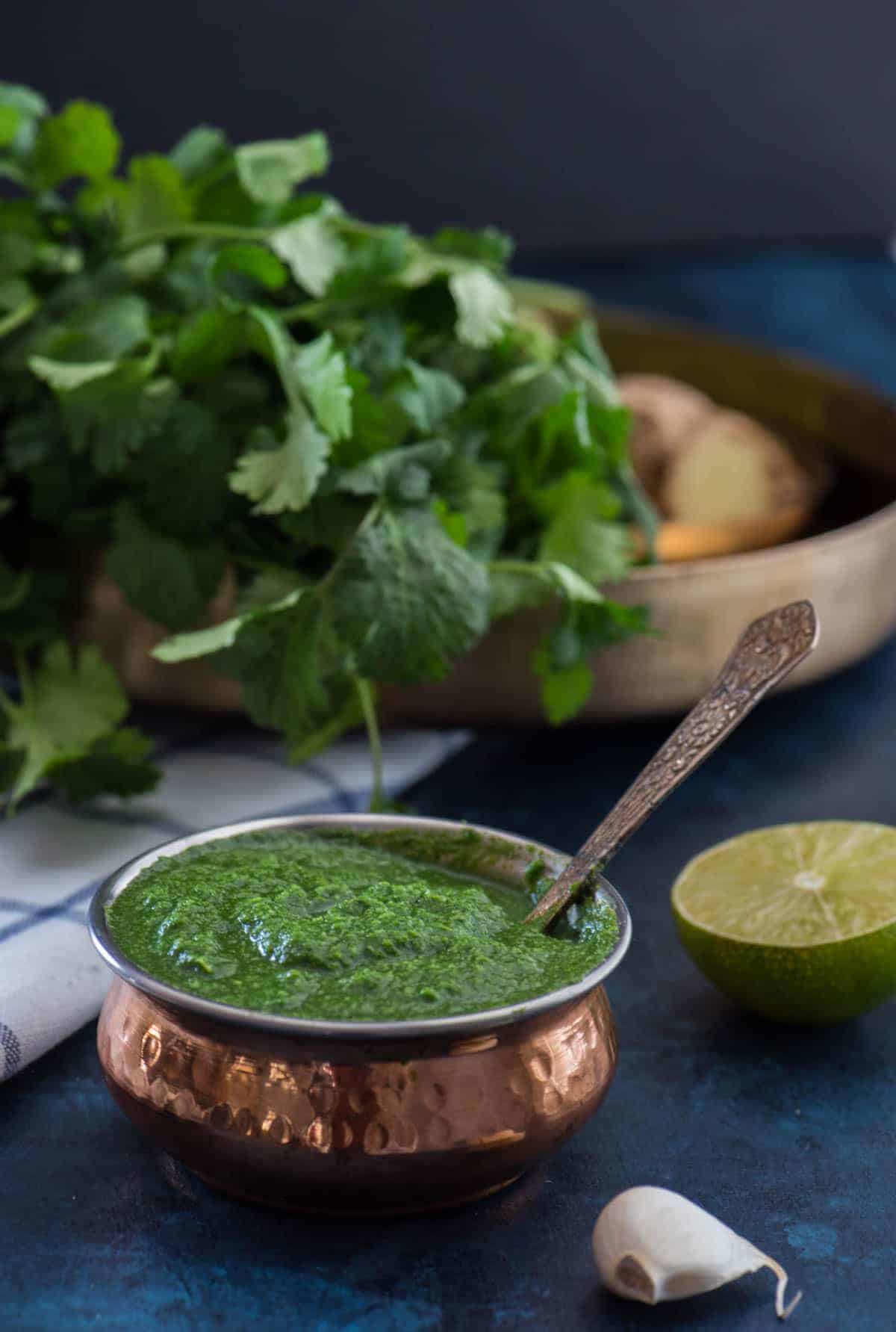 Green chutney served in a copper bowl. Also shown are the ingredients for coriander chutney - 1/2 of lime, garlic, coriander/cilantro leaves