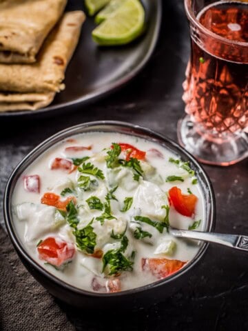 Cucumber tomato raita served in a black bowl and is accompanied by rotis and a glass of raspberry lemonade