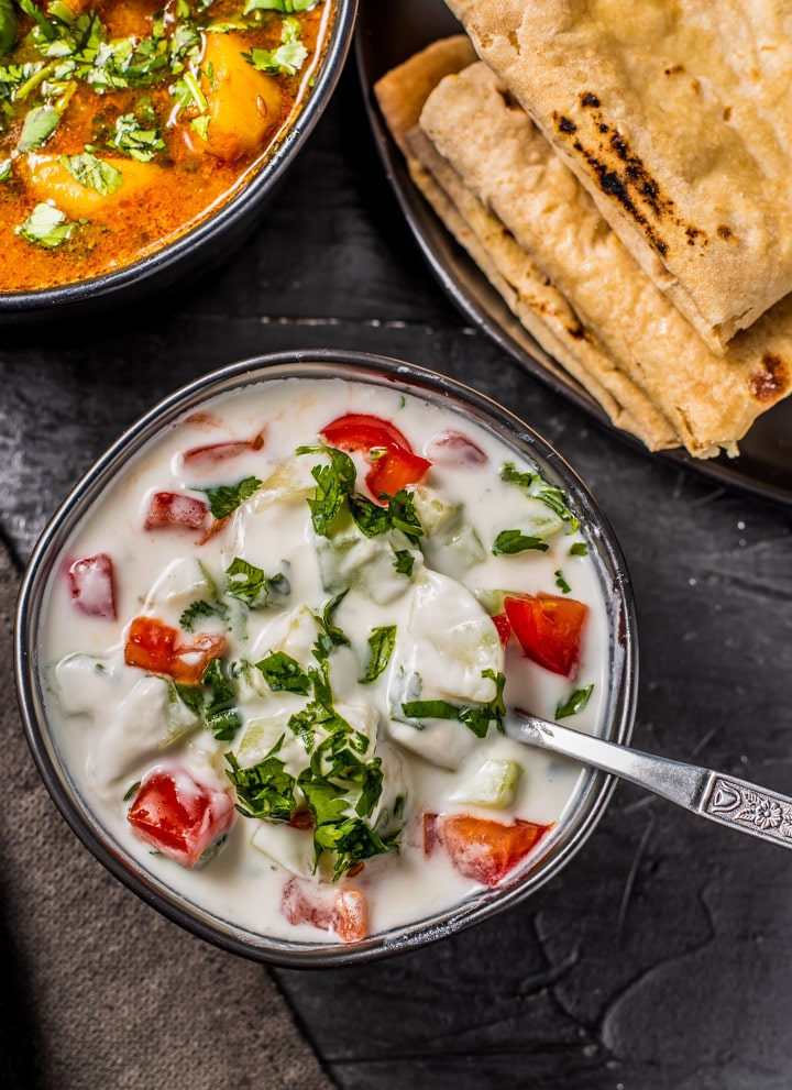 Cucumber tomato raita served in a black bowl and is accompanied by rotis and a bowl of potato curry