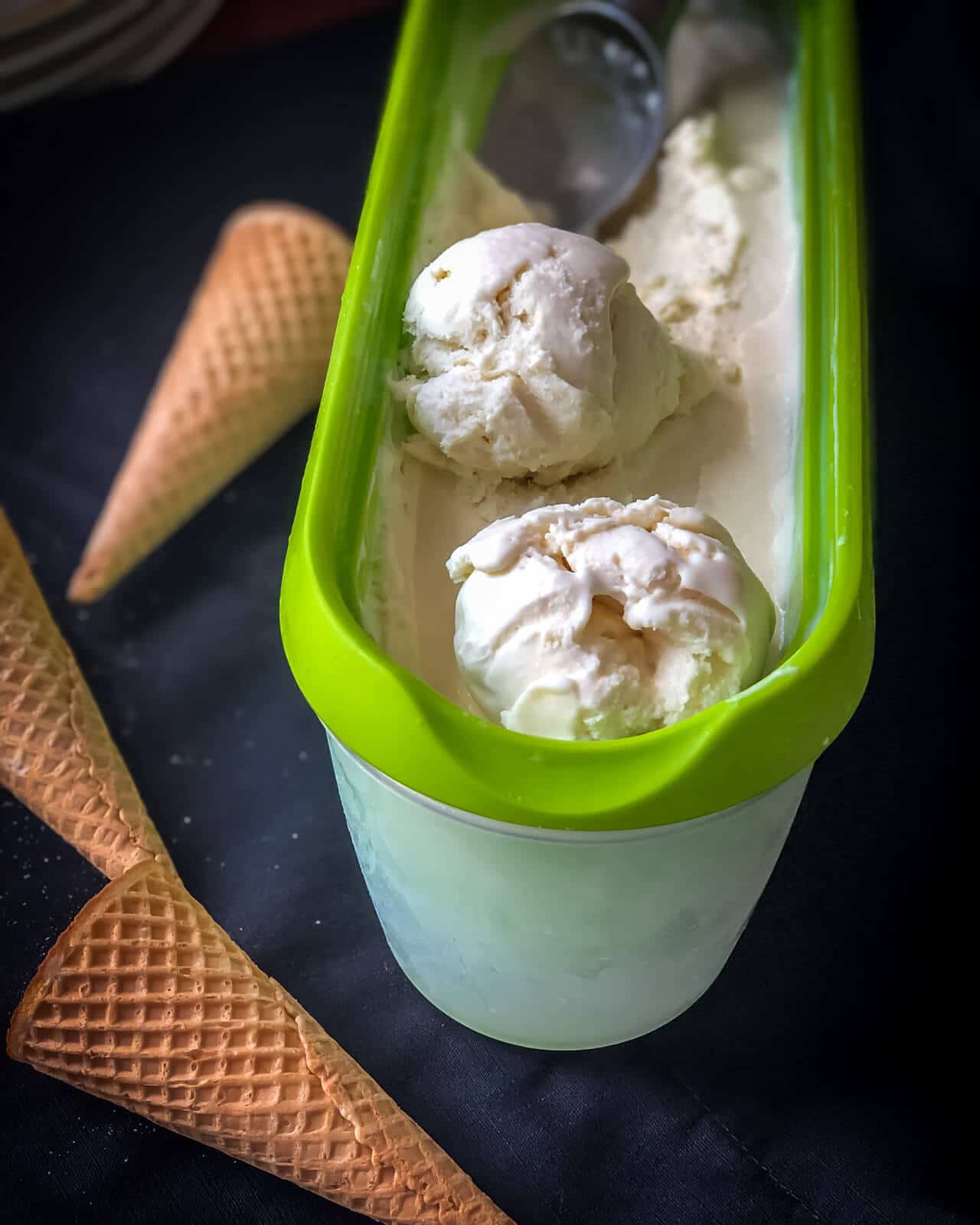 Vanilla ice cream in a tupperware with two rolled scoops and an ice cream scoop in the back and 3 ice cream cones on the right.