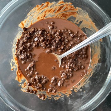 A glass bowl with almost fully melted chocolate chips and coconut oil stirred with a silver spoon.