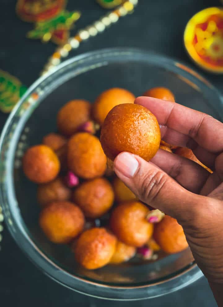 A glass bowl of milk powder gulab jamun balls on a dark counter with a hand in front holding a gulab jamun.