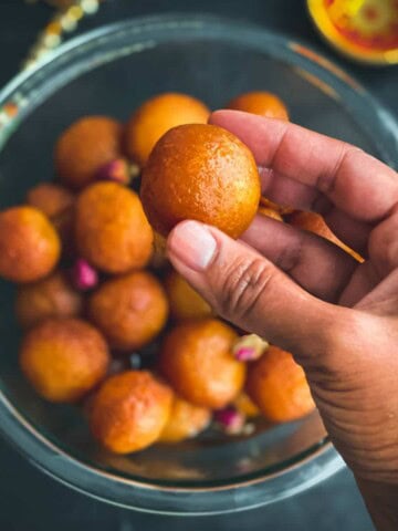 A hand holding a ball of gulab jamun over a glass bowl filled with gulab jamun balls.
