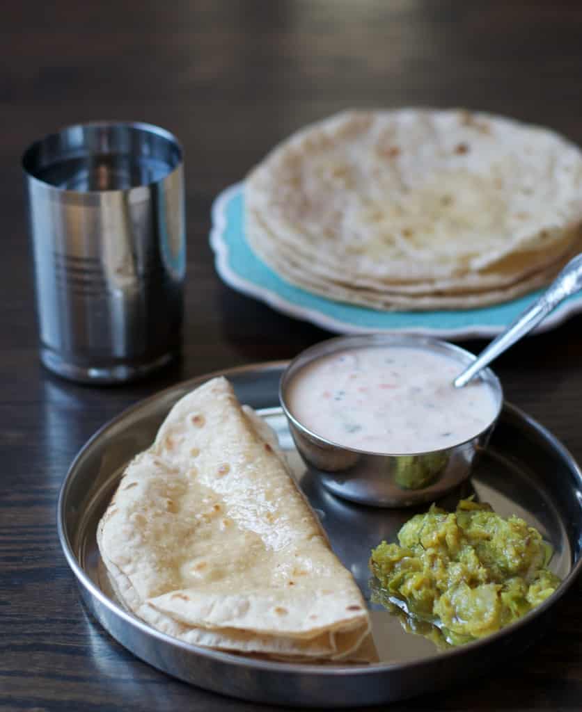 Ghee smeared soft rotis served with raita and a vegetable dish.