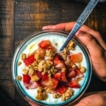 A hand holding a bowl of yogurt topped with strawberries, granola and honey