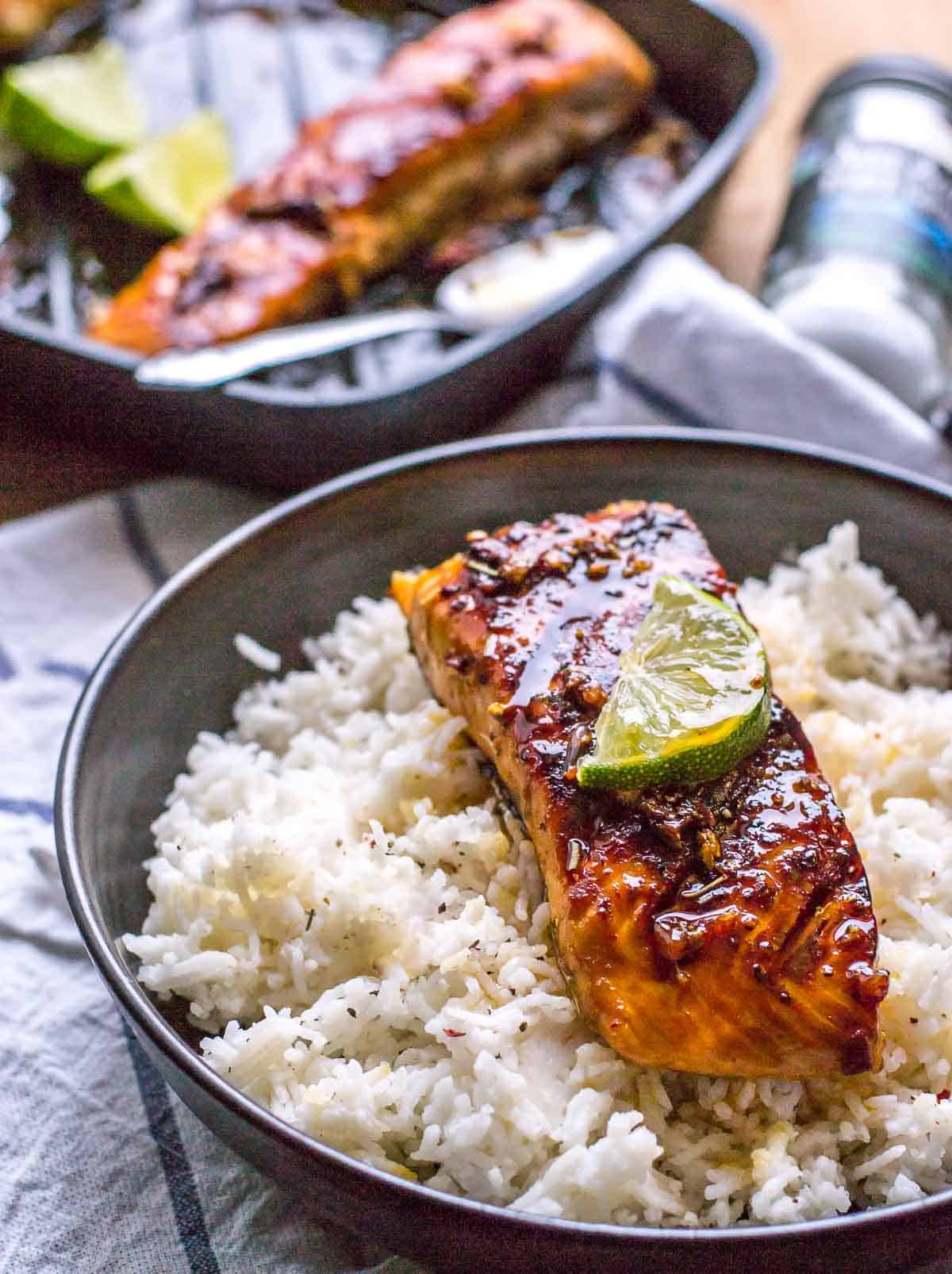 Honey garlic salmon with a lime slice on top served over a bed of rice in a black bowl with a bottle of spice and skillet on the side