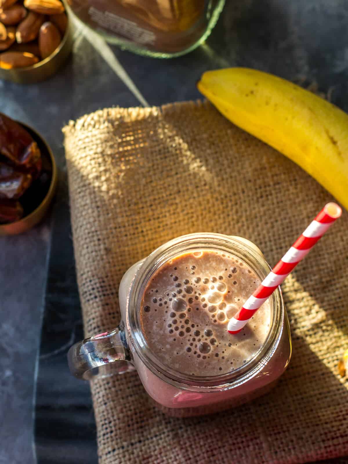 Chocolate almond smoothie served with a red and with straw. There is also a cocoa mix bottle along with almonds, dates and bananas in this picture.
