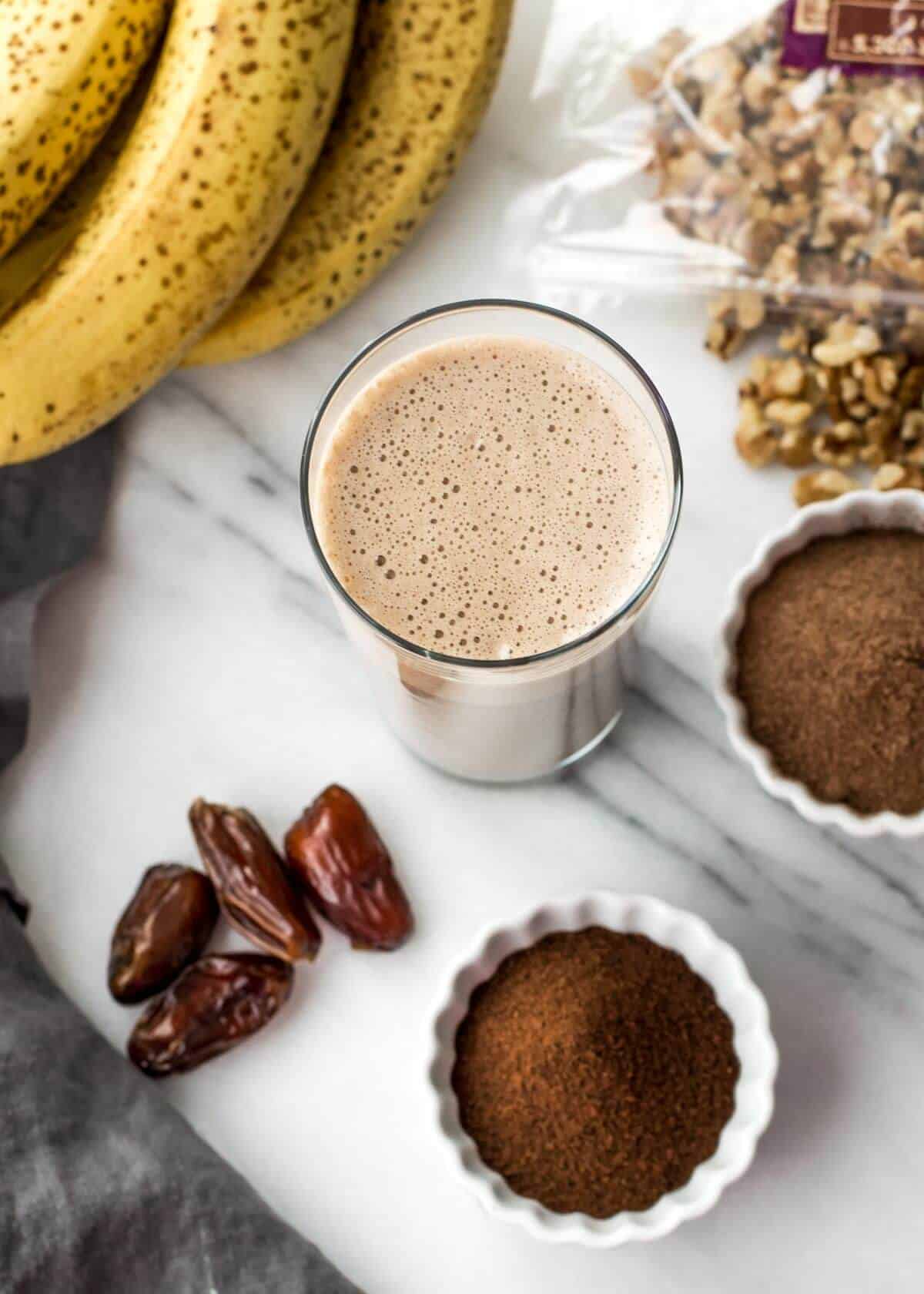 A glass of smoothie with banana. walnuts, coffee powder and dates next to it