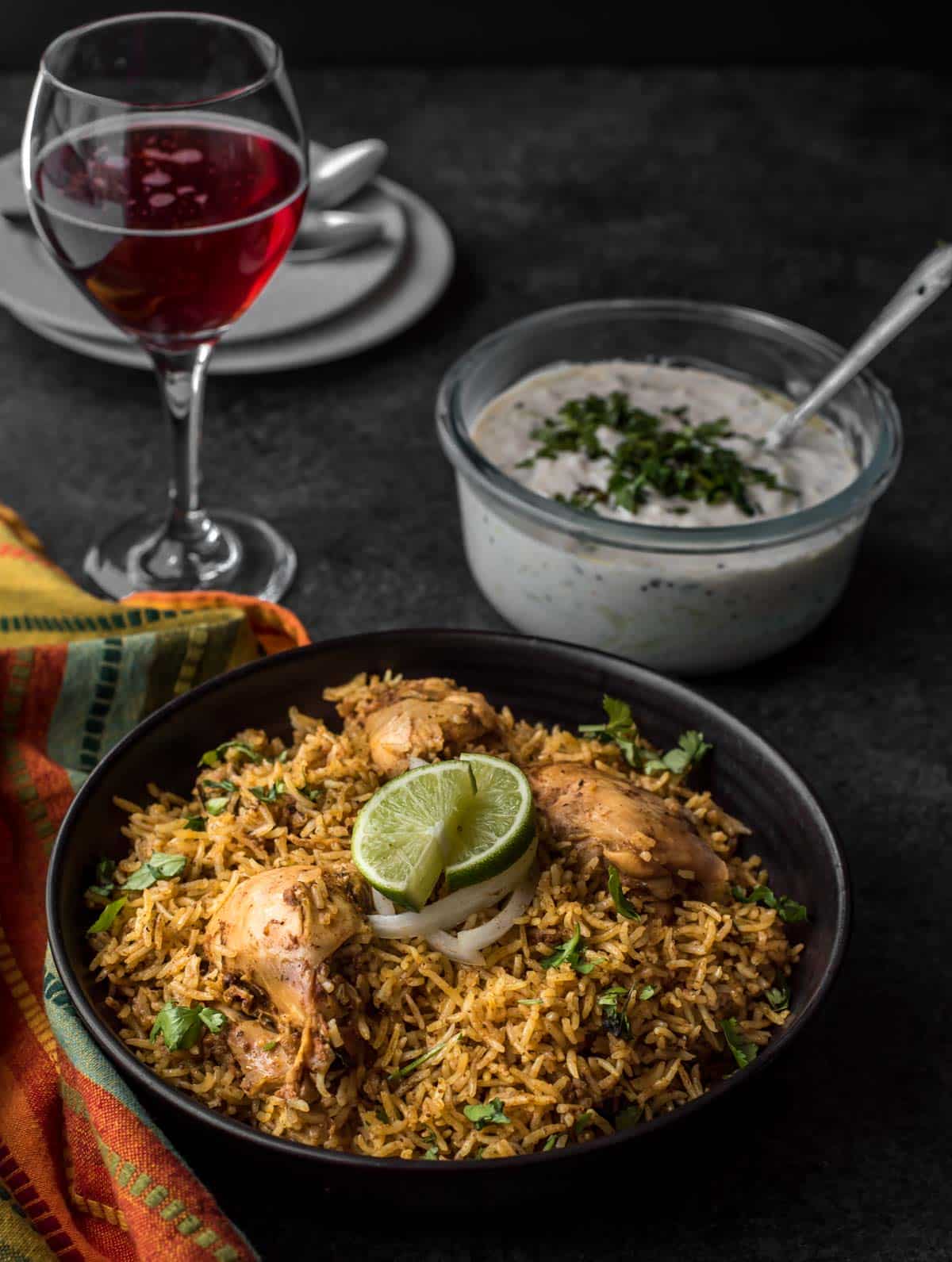 Chettinad Chicken biryani served in a black bowl with a side of raita and red sparkling wine