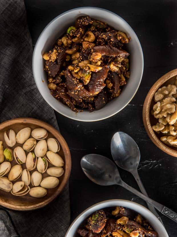 An overhead shot of fig and walnut halwa served in a white bowl with 2 spoons on the side. There are two other small wooden bowls filled with pistachios and walnuts respectively.