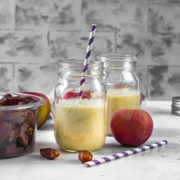2 glass jars filled with Apple smoothie. A box of dates, an apple, along with a purple straw are on the side.