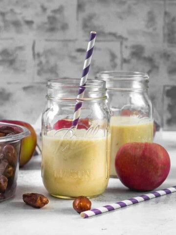 2 glass jars filled with Apple smoothie. A box of dates, an apple, along with a purple straw are on the side.
