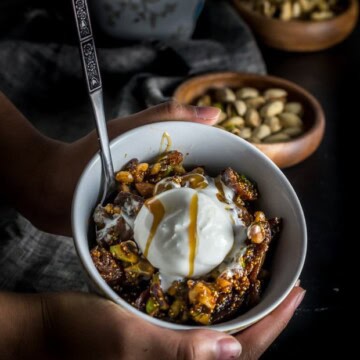 A hand holding a white bowl of fig and walnut halwa. A wooden bowl of pistachios is seen in the background.