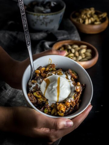 A hand holding a white bowl of fig and walnut halwa. A wooden bowl of pistachios is seen in the background.