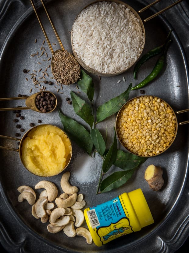 An overhead shot showing ingredients used in Ven Pongal Recipe. Includes cashews, curry leaves, salt, ginger, moong dal, rice, cumin seeds, pepper, green chilies and asafoetida. 