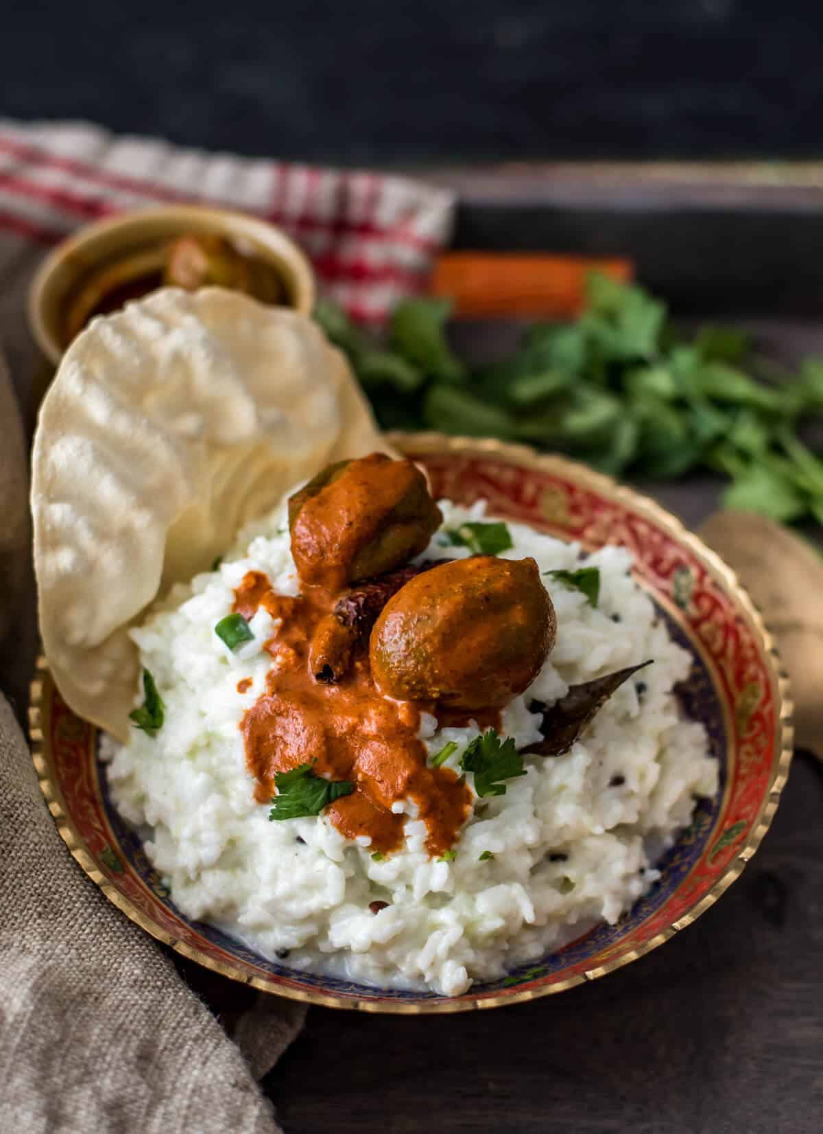 Curd rice served in a metallic gold and red bowl topped with mango pickle and papad. It is also accompanied by pickle in a ceramic brown bowl with coriander on the side