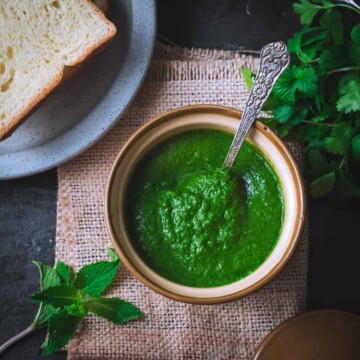 An overhead shot of coriander mint chutney placed in a porcelain bowl. There is a grey plate with bread slices, a bunch of cilantro and a few mint leaves on the side.