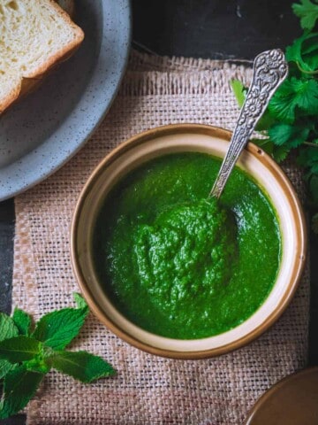 An overhead shot of coriander mint chutney placed in a porcelain bowl. There is a grey plate with bread slices, a bunch of cilantro and a few mint leaves on the side.
