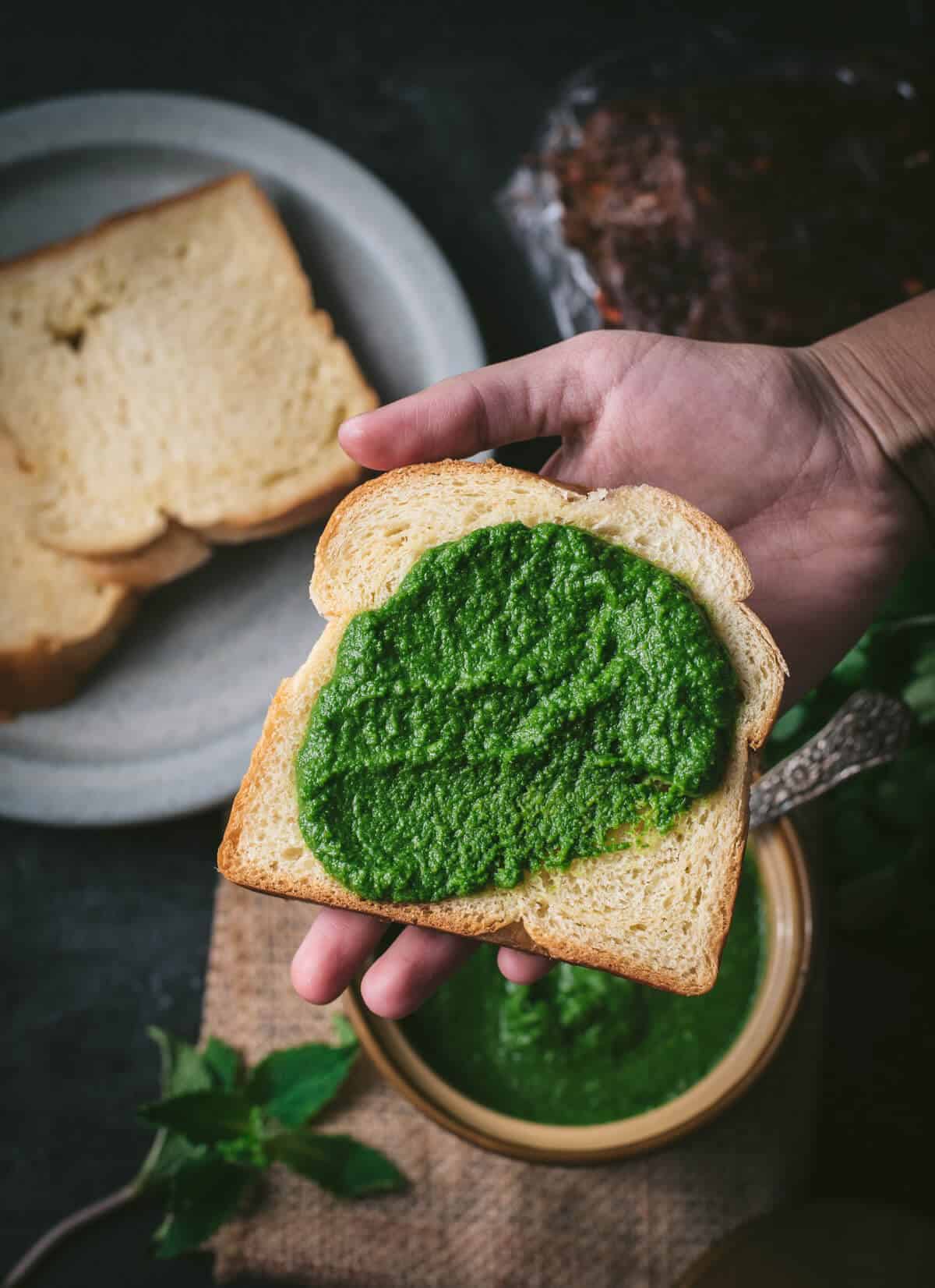 Bread slathered with green chutney is placed on a palm. There are additional bread slices kept on a grey plate. 