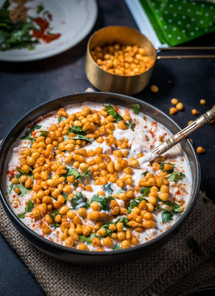 Boondi raita recipe is placed on a black bowl with a golden spoon. A measuring cup holds boondi raita while a glass bowl holds remnants of boondi raita.