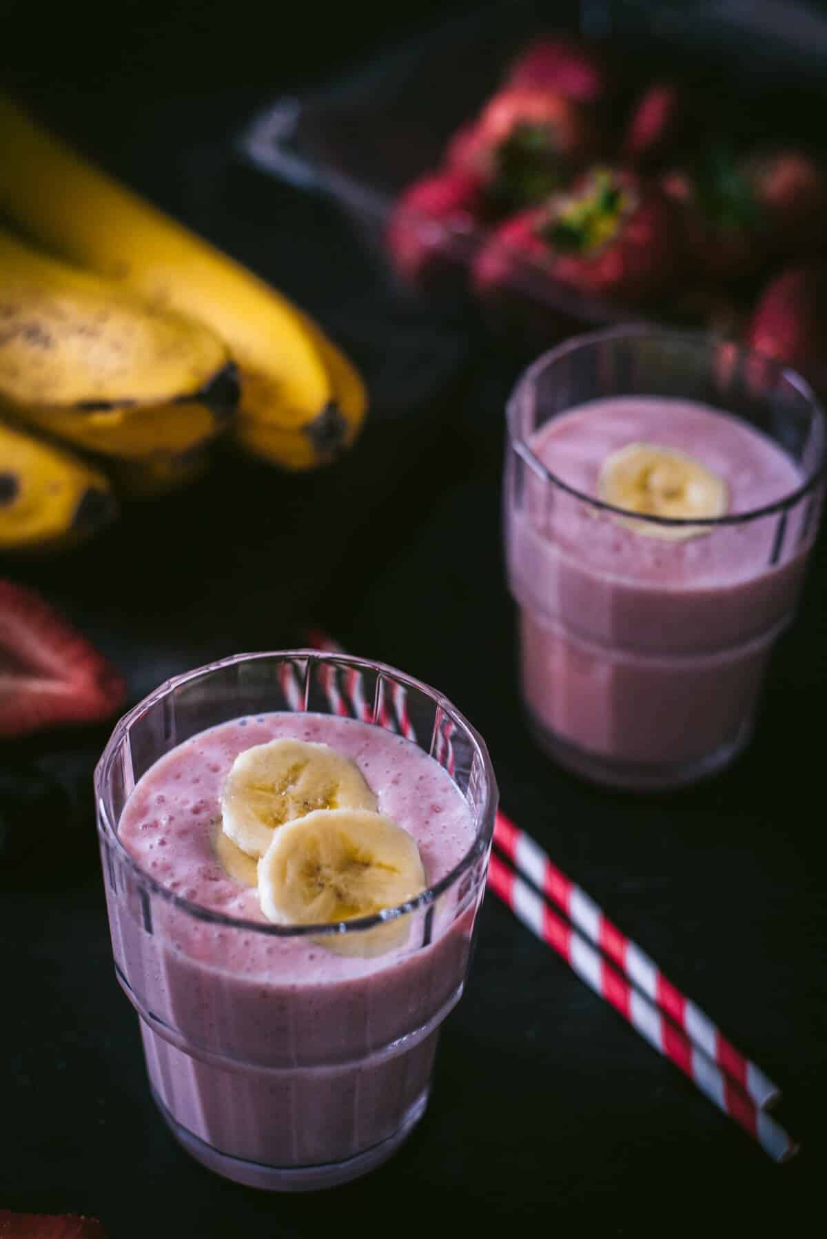 An overhead shot of strawberry banana smoothie in two glass containers and placed on a wooden bowl. There are two red and white straws on the side.
