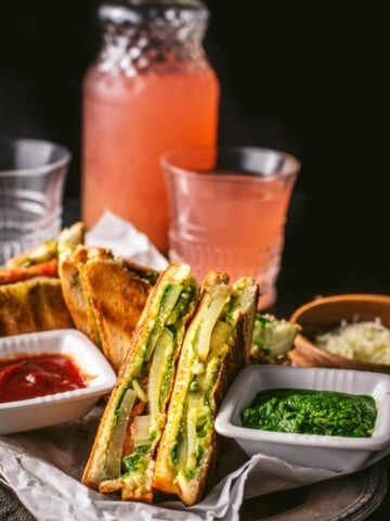 Bombay Veggie Grilled Sandwich served with a side of green chutney and ketchup. A bottle and a glass of raspberry lemonade is also placed on the side along with grated cheese