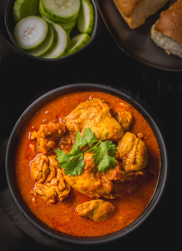 An overhead shot of Chicken Vindaloo served in a black bowl. Served with cucumber slices and pav (dinner rolls)