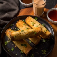 An overhead shot of bread rolls served with tamarind chutney and ketchup (served in white bowls) and a glass of tea