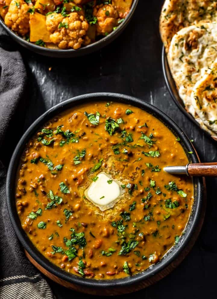 An overhead shot of Dal Makhani recipe served with a sliver of butter and accompanied with garlic naan and aloo gobi