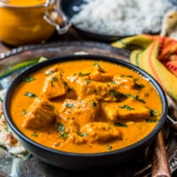A black bowl of salmon tikka masala on a silver platter with naan, masala sauce, and rice in the background.