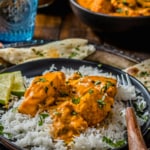 Chunks of salmon tikka masala are served over a bed of rice with lime wedges on a black plate. This plate sits on a silver platter with naan, a cup of water, and a bowl of salmon tikka masala in the background.