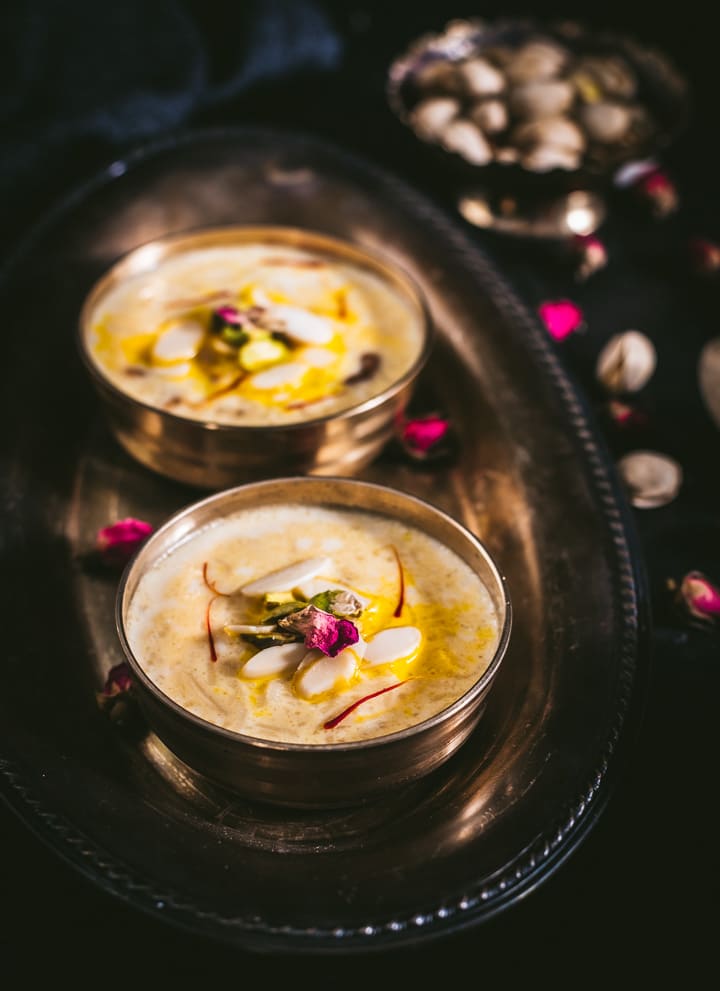 Two small bowls of creamy rice kheer on a silver platter on the left of the picture with mixed nuts in a small bowl of the right of the picture all on a black table cloth with almond slivers scattered on top.