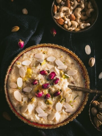 A large bowl of Rice Kheer in a gold rimmed bowl with almond slices and a spoon on top in the bottom left of and a bowl of mixed nuts in the top right on a black table cloth with almond slivers scattered around.
