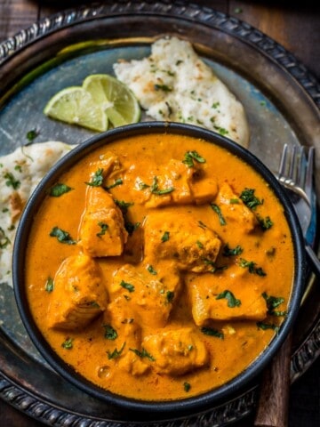 A black rimmed bowl of salmon curry on a silver platter with naan, lime wedges, and a wooden handled fork.