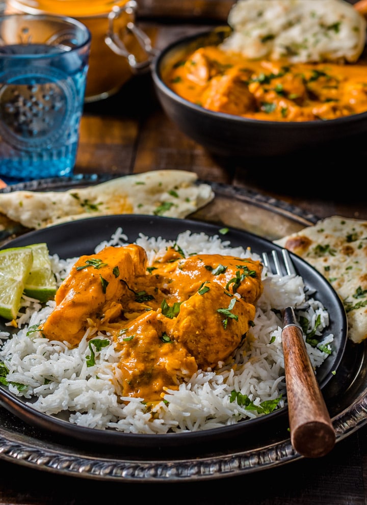 Chunks of salmon tikka masala are served over a bed of rice with lime wedges on a black plate. This plate sits on a silver platter with naan, a cup of water, and a bowl of salmon tikka masala in the background.