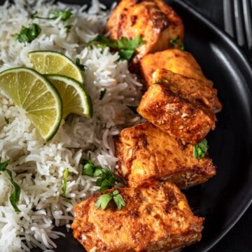 Full plate of Tandoori Salmon cooked to perfection and served on a black plate with cooked rice and garnished with lime and cilantro.