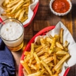 Two red baskets of french fries with parchment paper, a denim napkin, beer, and a cup of ketchup on a wooden table with the words Homemade Air Fryed French Fries on the top.
