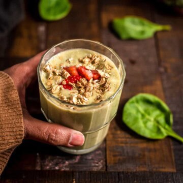 A hand holding a glass of strawberry banana spinach smoothie garnished with strawberry and granola