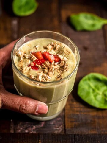 A hand holding a glass of strawberry banana spinach smoothie garnished with strawberry and granola