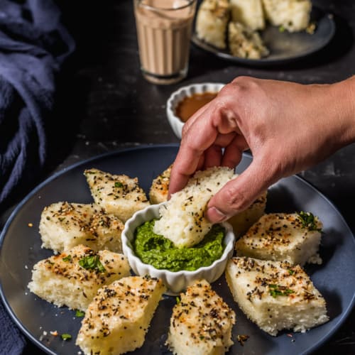 A hand dipping a piece of rava dhokla in chutney.