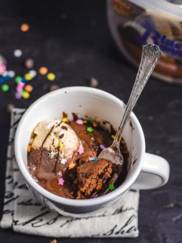 A white coffee mug with a chocolate mug cake inside topped with vanilla chocolate ice cream, sprinkles and chocolate sauce with a fork sticking out of the mug.