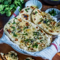 A fresh stack of garlic naan with chopped cilantro on a white dish towel next to a cup of cilantro.