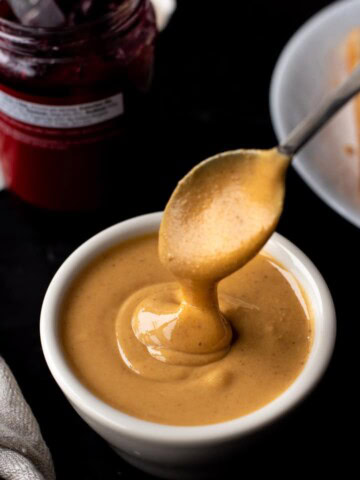 A white bowl of creamy peanut butter with a spoon scooped in the peanut butter.